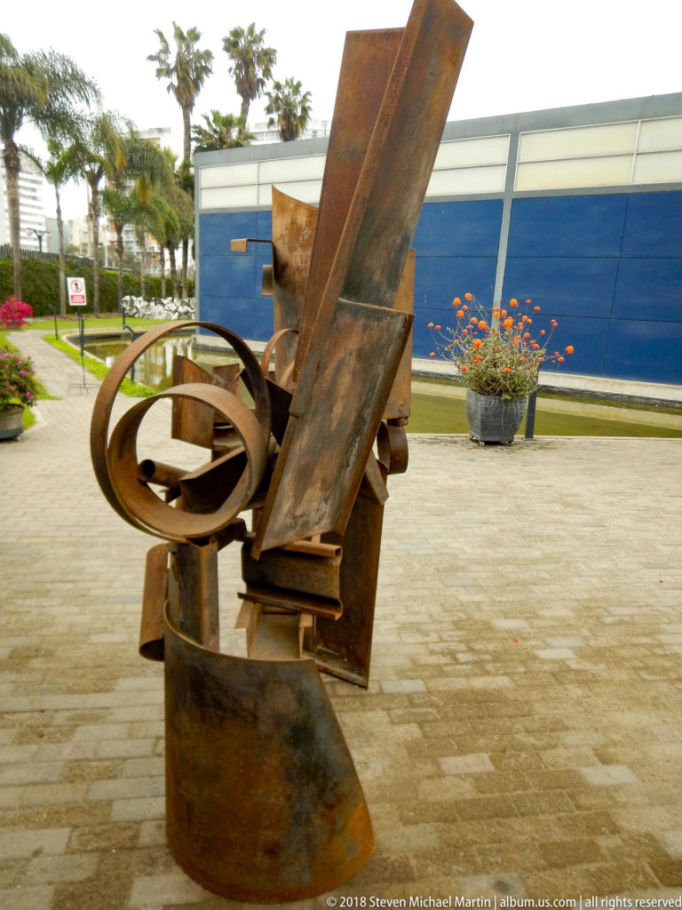 Large rusted-steel abstract sculpture with intersecting plates and a circular element standing in a paved museum courtyard.