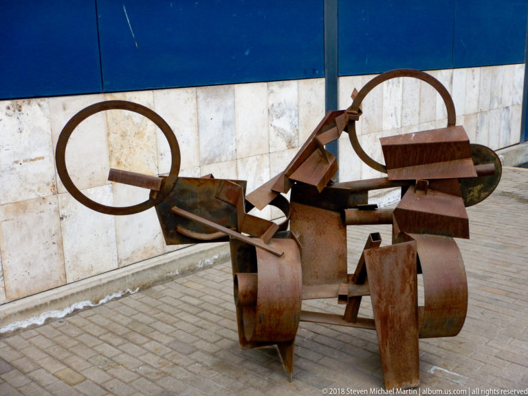 Rusted welded-steel abstract sculpture made from rings, wheels, curved plates, beams, and blocklike forms.