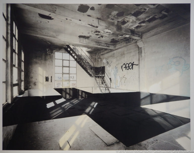 Angular black floor bands crossing a distressed room toward a stair and sunlit windows.