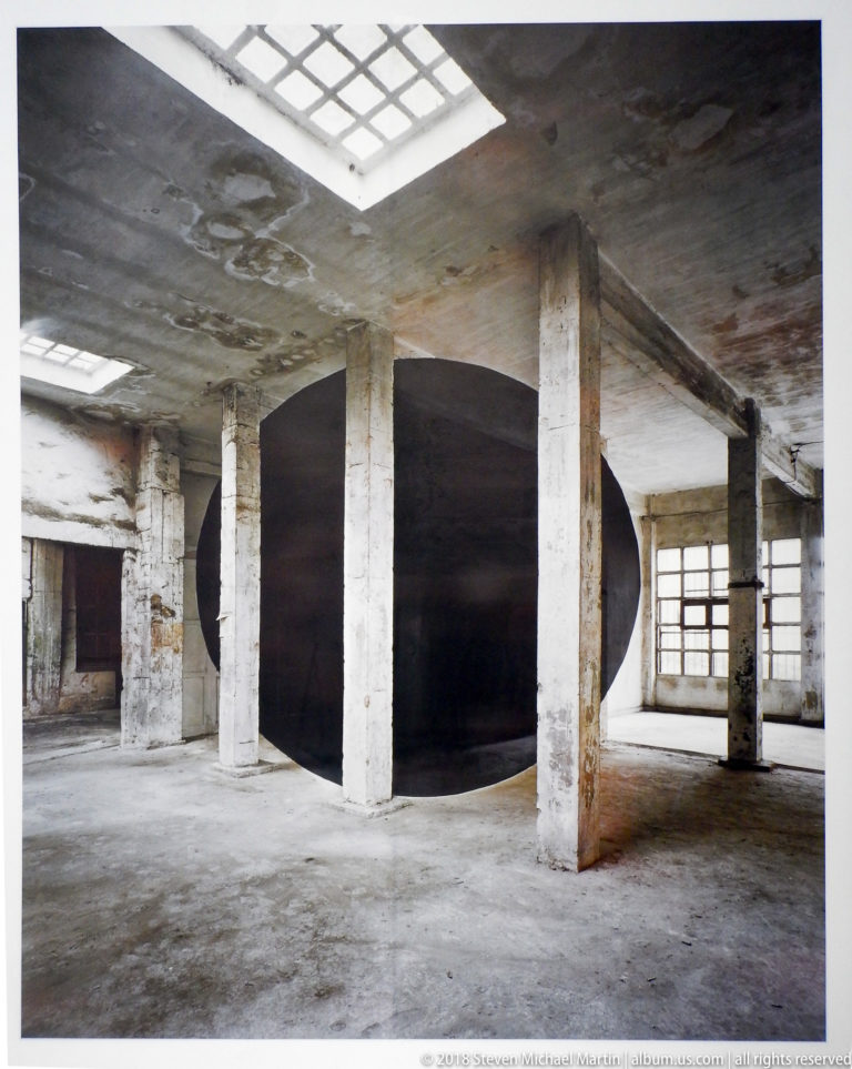Large matte-black horizontal cylindrical installation between columns in a raw concrete room.