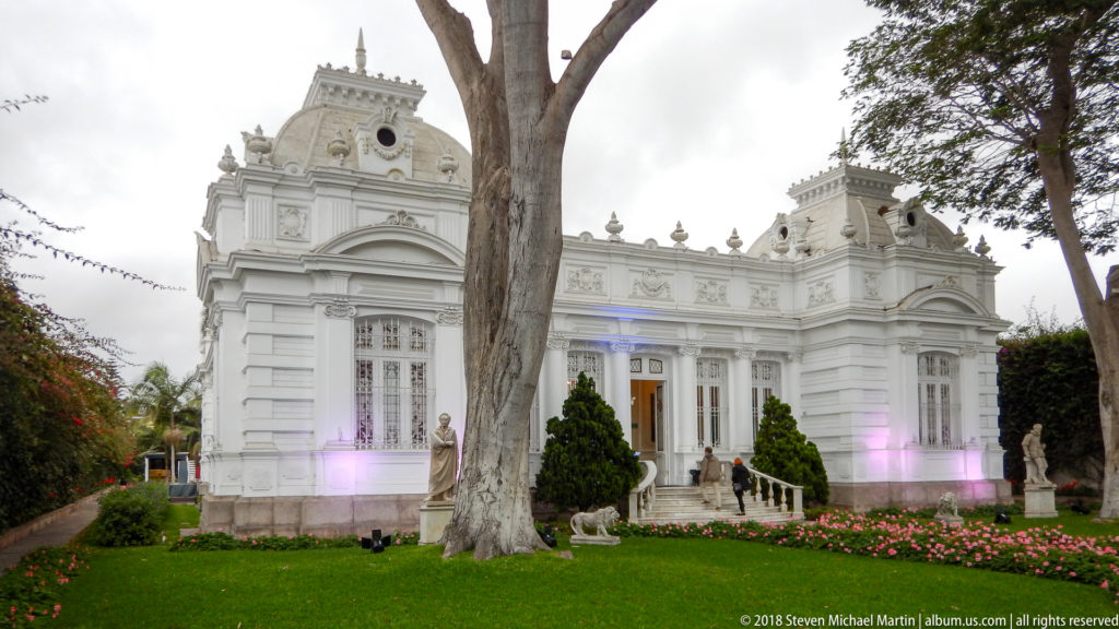 White ornate Pedro de Osma Museum mansion behind a lawn, flower beds, trees, sculptures, and colored façade lighting.