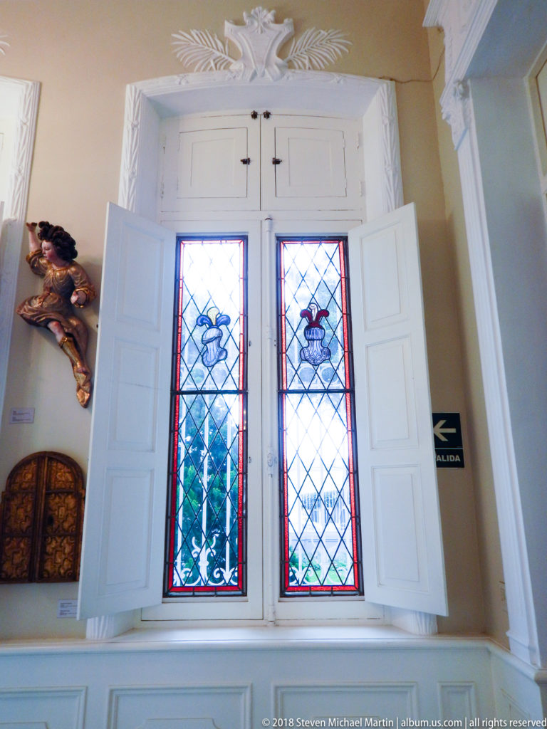Two narrow stained-glass windows with stylized bird motifs, open white shutters, plaster ornament, and a carved figure nearby.