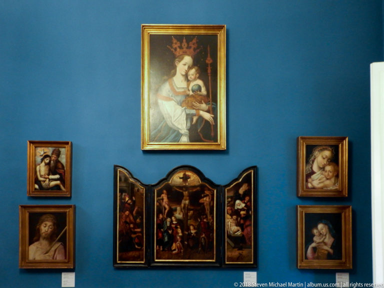 Blue museum wall displaying a crowned Virgin and Child, a small Crucifixion triptych, and several smaller devotional paintings.