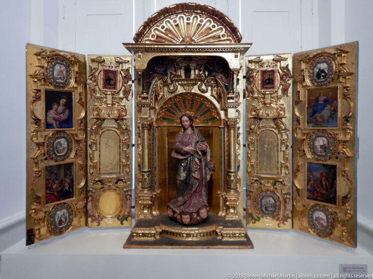 Open gilded devotional shrine with central female saint sculpture, painted scenes on hinged wings, and dense architectural ornament.