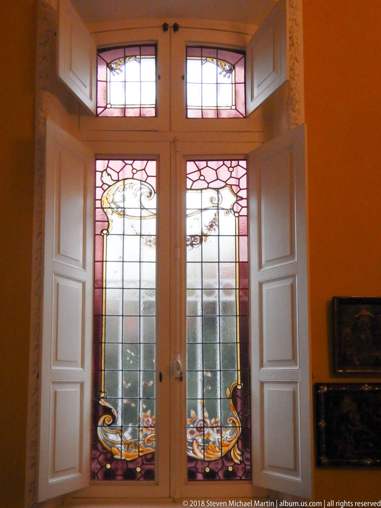 Paired stained-glass doors with pink borders and floral scroll motifs, framed by open white shutters in a warm-toned room.