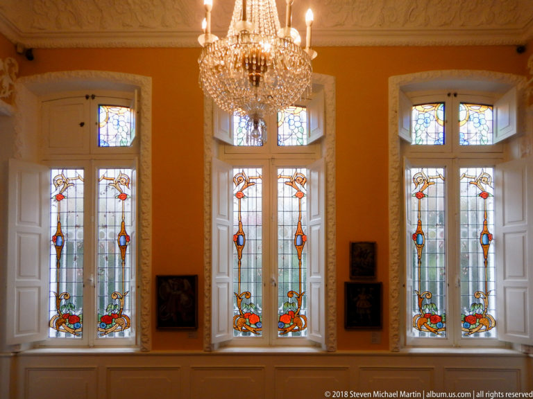 Three tall stained-glass window groups with repeated colorful motifs, open white shutters, and a crystal chandelier overhead.