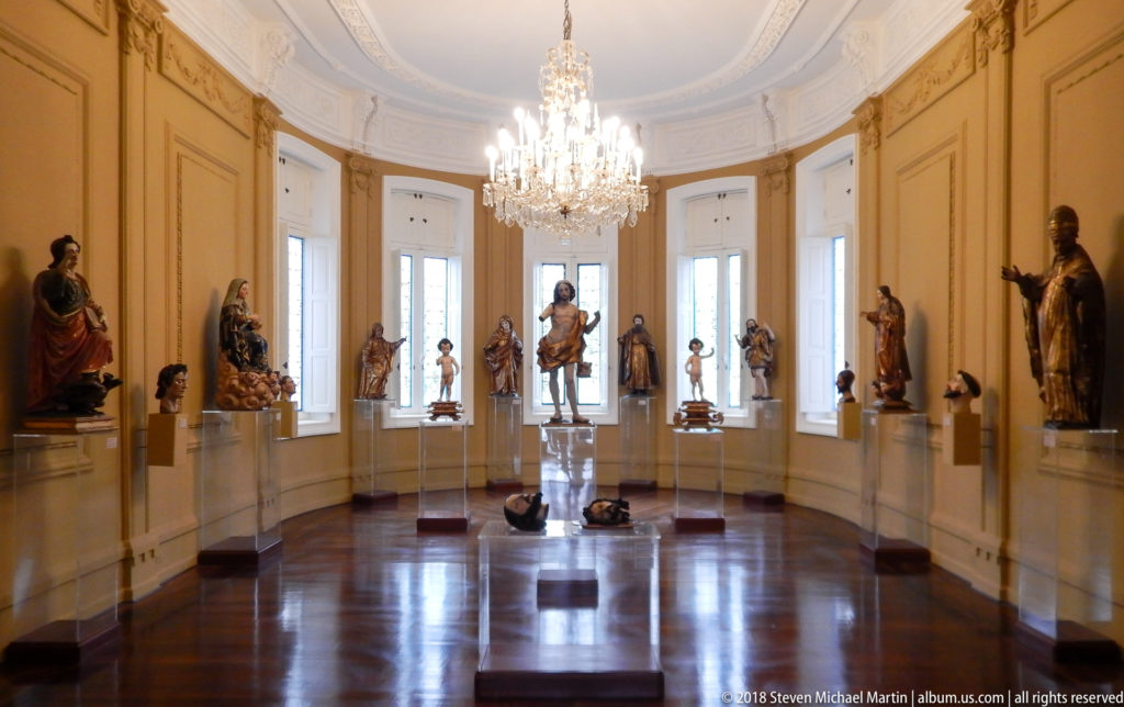 Curved museum gallery lined with polychrome religious sculptures, tall windows, polished parquet floor, and a crystal chandelier.