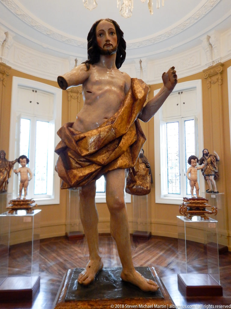 Polychrome standing Christ figure with extended arms and gold-toned mantle, with other religious sculptures displayed behind.