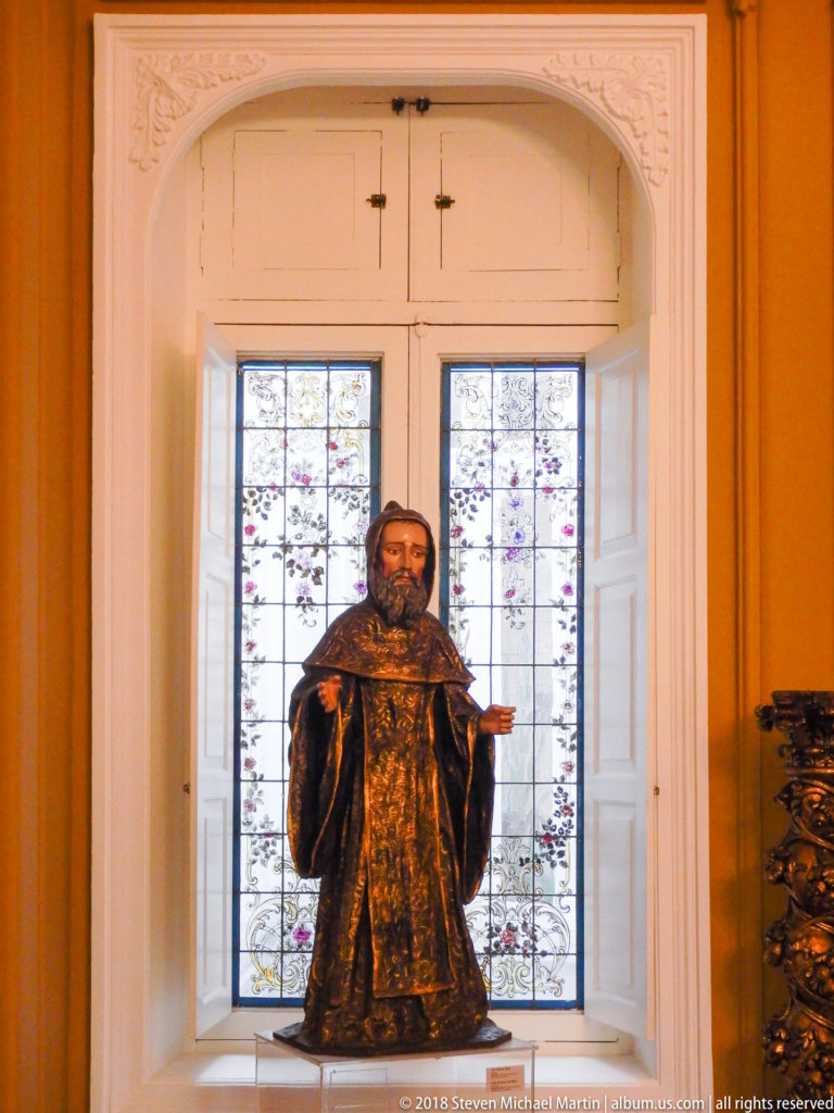Standing bearded saint sculpture in a dark gold-patterned robe, displayed against a tall window with open white shutters.