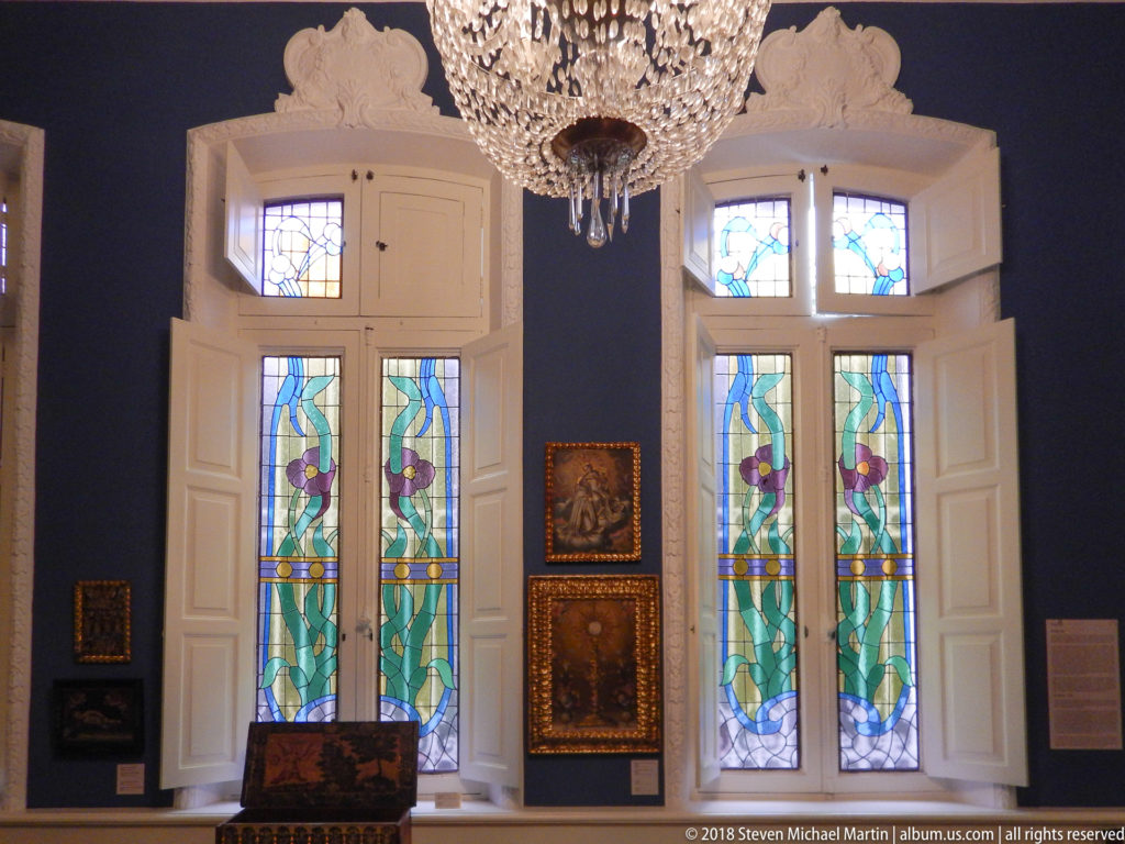 Tall floral stained-glass windows with white shutters, surrounded by small framed paintings and a decorated chest.