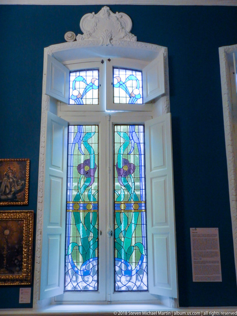 Tall paired stained-glass window with curving green, blue, and amber motifs, framed by open white shutters.
