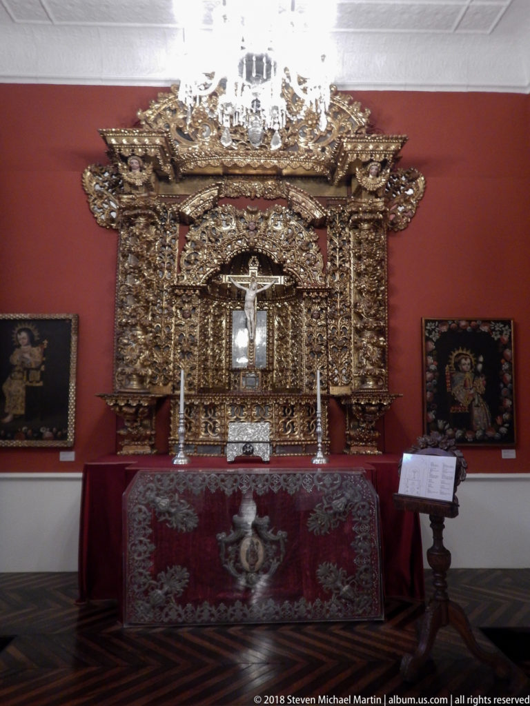 Gilded chapel-style retable with central crucifix above a red embroidered altar frontal, flanked by devotional paintings.