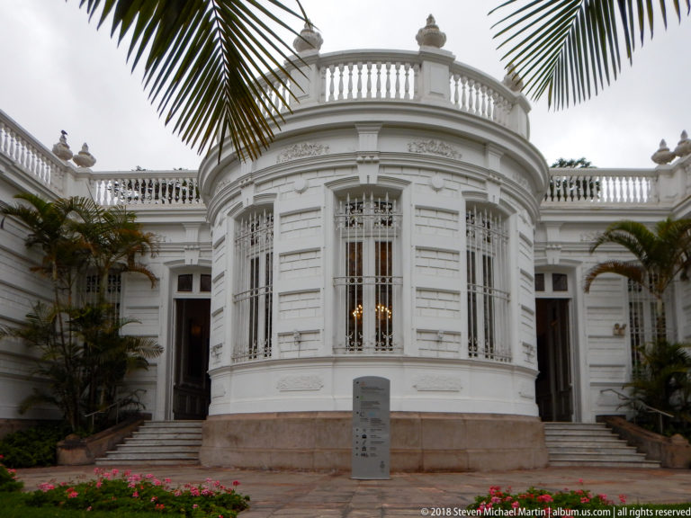 Rounded white bay with tall grilled windows, curved roof balustrade, side stairs, palms, and flower beds.