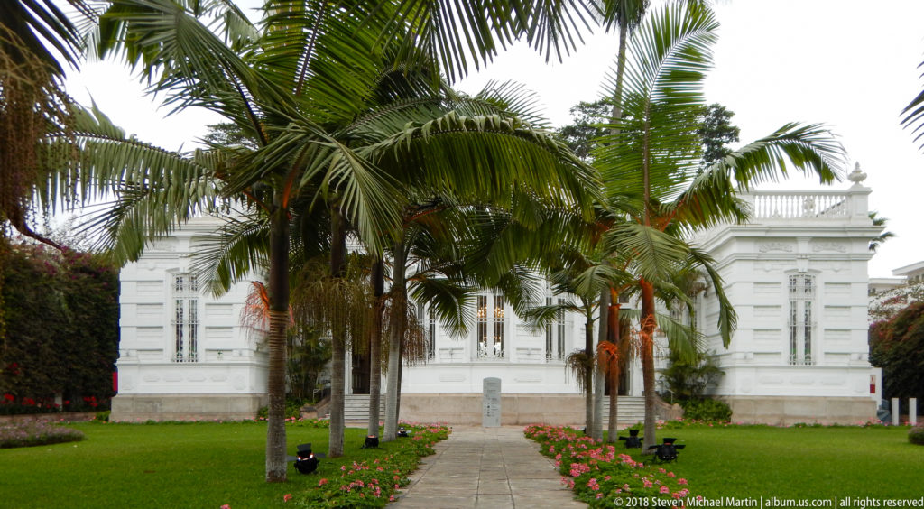 Palm-lined garden path with pink flower borders leading toward the white Pedro de Osma Museum mansion.