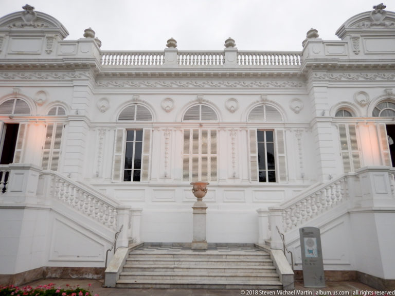 White historic mansion façade with tall shuttered windows, decorative moldings, roof balustrade, and symmetrical exterior stairways.