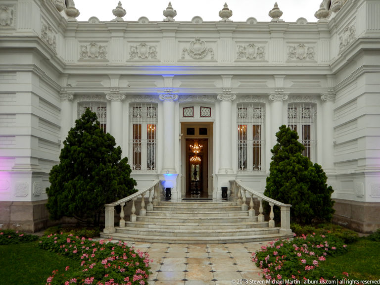 Central white museum entrance with steps and curved balustrades, tall grilled windows, clipped shrubs, and pink flower beds.