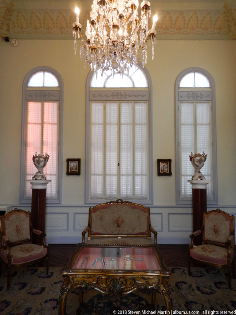Formal salon with crystal chandelier, upholstered seating, central table, arched windows, and matching pedestal-mounted decorative vessels.
