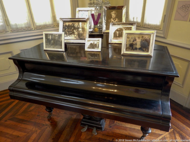 Black grand piano topped with numerous framed historic photographs in a bright room with parquet flooring.