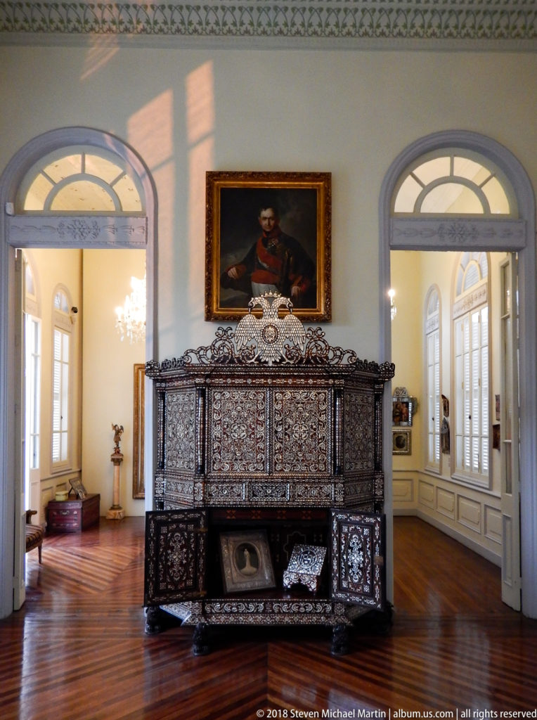 Frontal view of a broad ornate inlaid cabinet beneath a uniformed man’s portrait, flanked by tall arched openings.