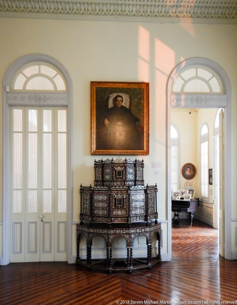 Inlaid cabinet centered beneath a portrait between two tall arched doorways, with sunlight crossing the wall and parquet floor.