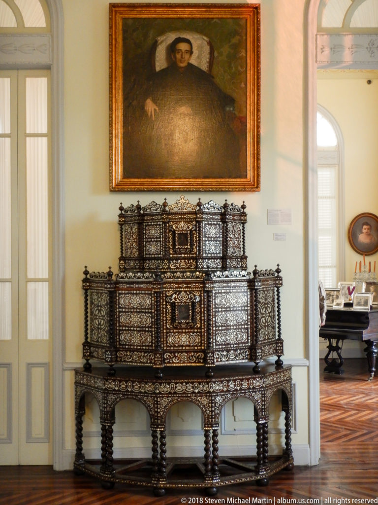 Tiered dark-wood inlaid cabinet on an arcaded stand beneath a male portrait, with an adjoining gallery visible at right.