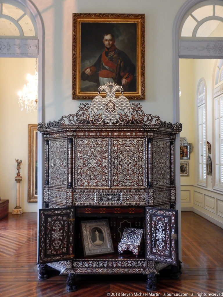 Large dark inlaid cabinet with open lower doors beneath a framed male portrait in a bright museum gallery.