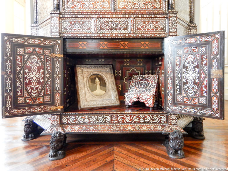 Open inlaid wooden cabinet with geometric marquetry, carved feet, framed devotional image, and decorated textile inside.