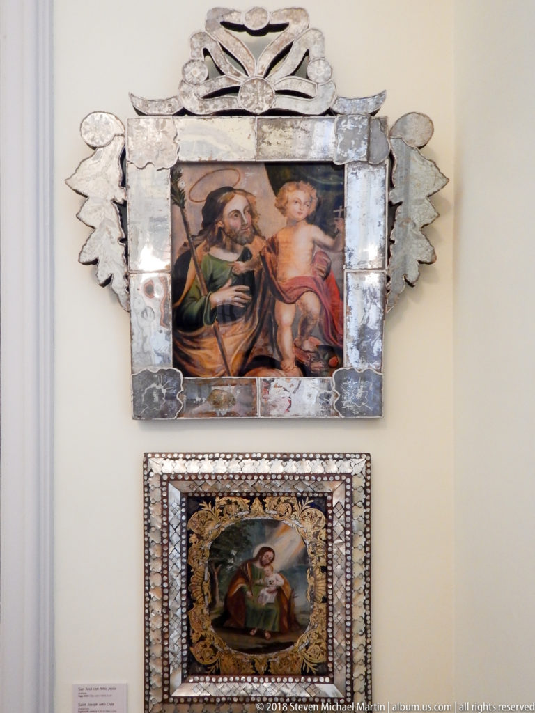 Two small religious paintings mounted vertically, the upper in a silver-toned radiating frame and the lower in geometric inlay.