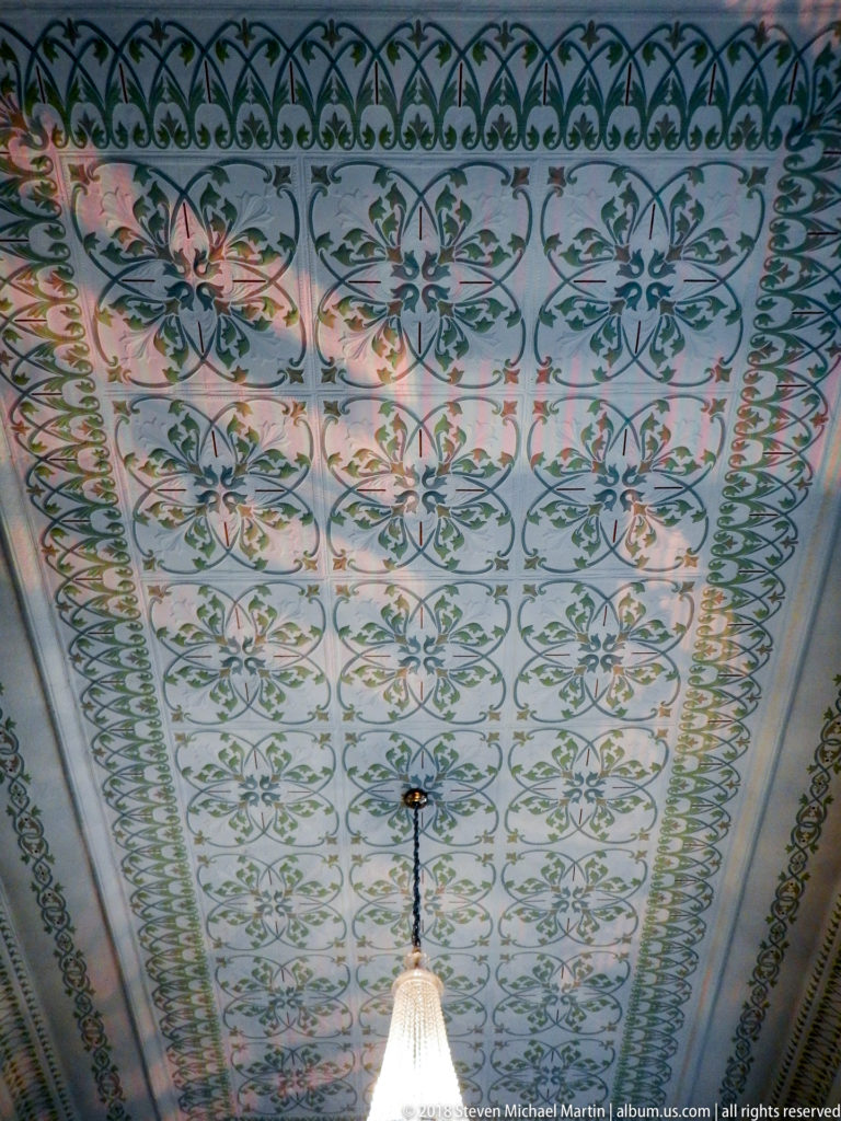 Painted ceiling covered with repeating green scrollwork and red floral motifs, centered on a hanging light fixture.