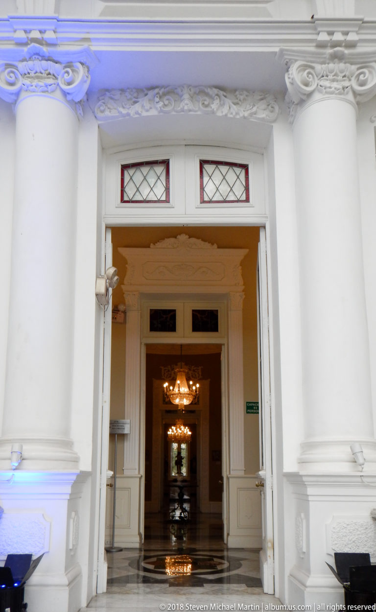 White molded museum doorway with leaded-glass transoms opening toward chandelier-lit rooms along a central axis.