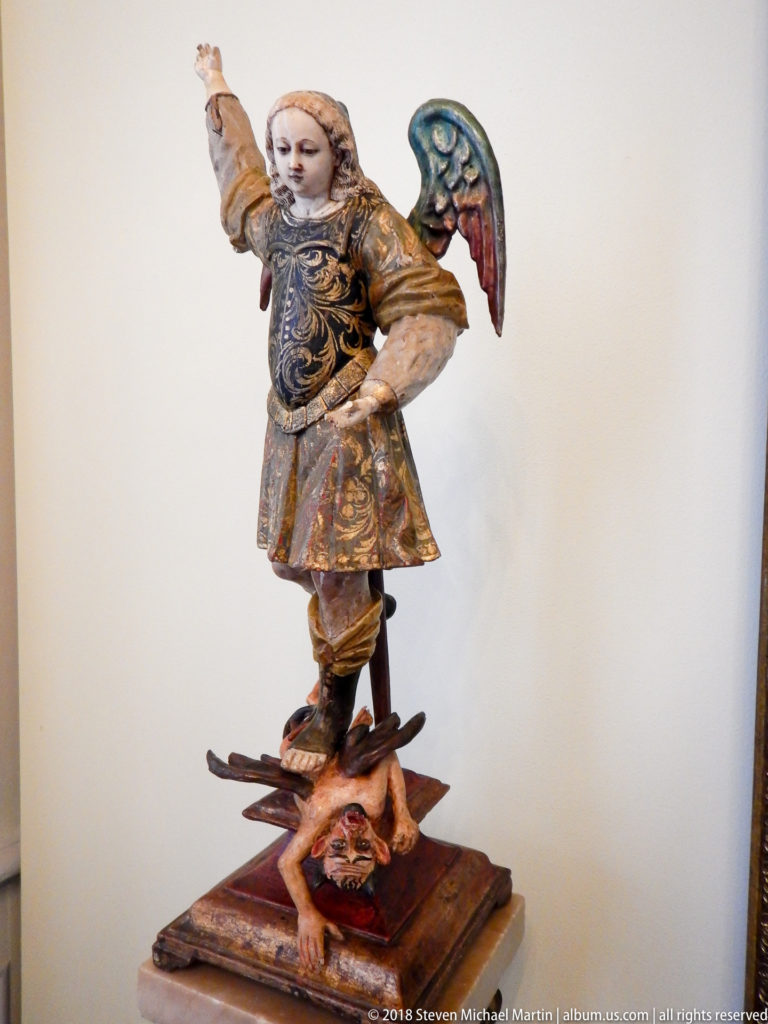 Polychrome carved archangel in patterned armor and wings standing over a defeated demon on a wooden base.