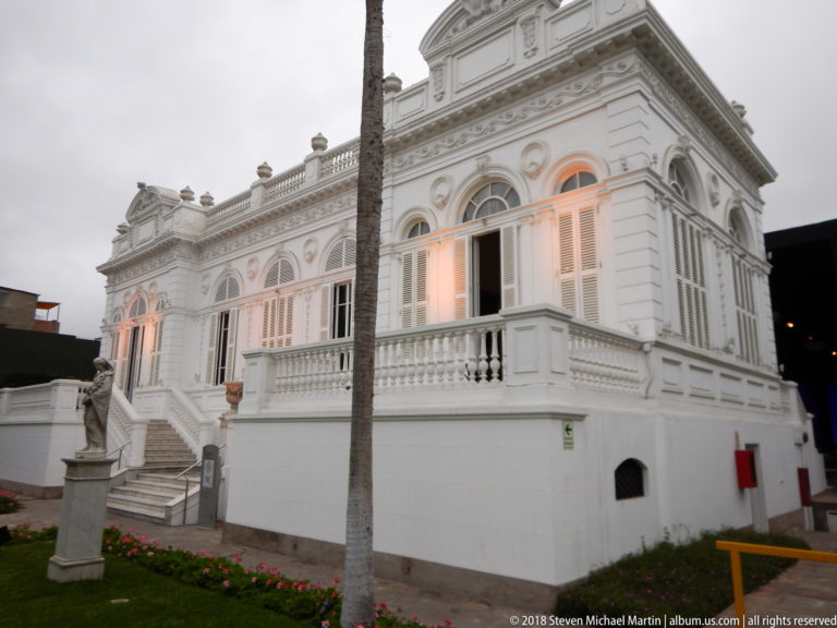 Oblique view of the white museum mansion showing exterior steps, projecting corner bay, arched windows, balustrade, and palm.