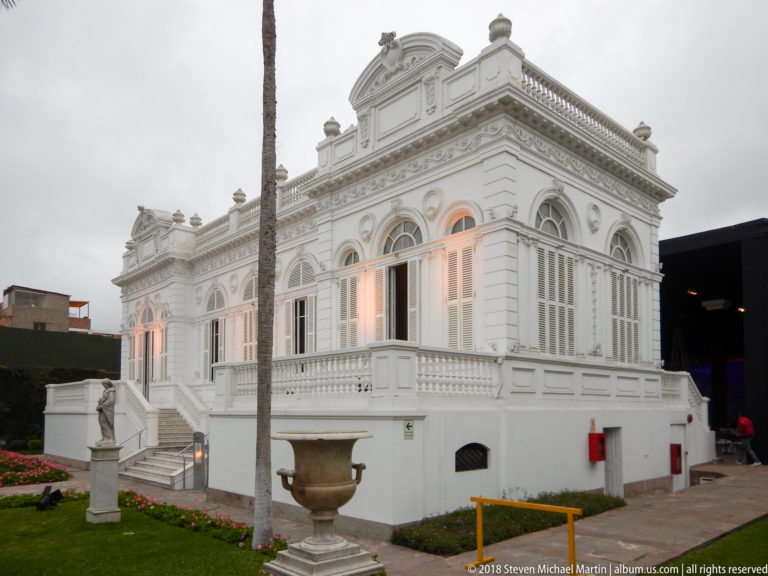 White corner pavilion of the museum with arched windows, terrace balustrade, palm trunk, stone urn, and garden sculpture.