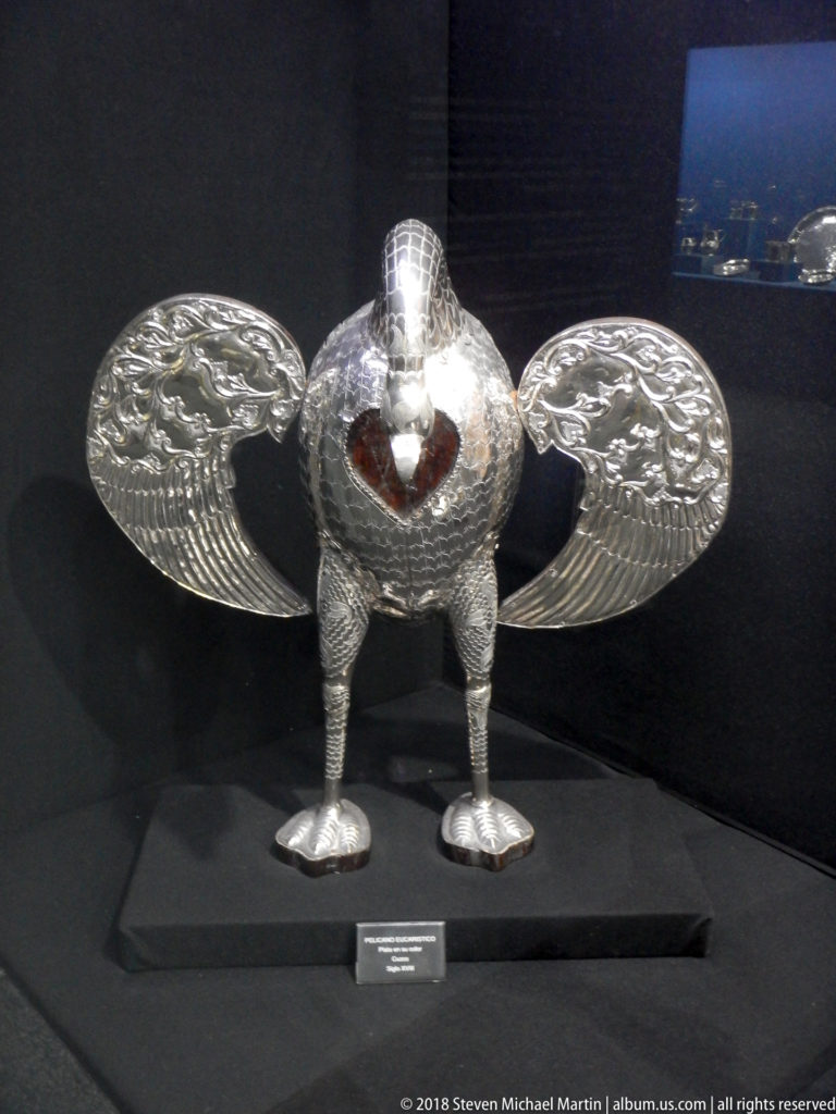 Winged silver bird-like devotional object with embossed feathers, two legs, and a red heart visible at the center.