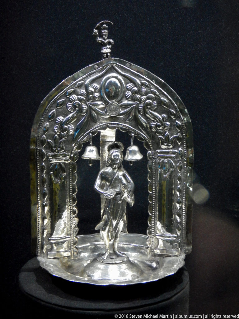 Small arched silver shrine containing a crucifixion scene, standing figures, columns, and embossed architectural ornament.