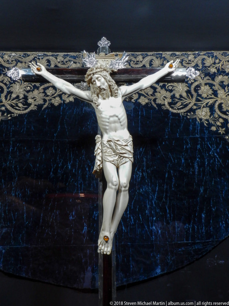 Pale sculpted Christ on a dark crucifix set against deep blue textile embroidered with gold scrolling foliage.