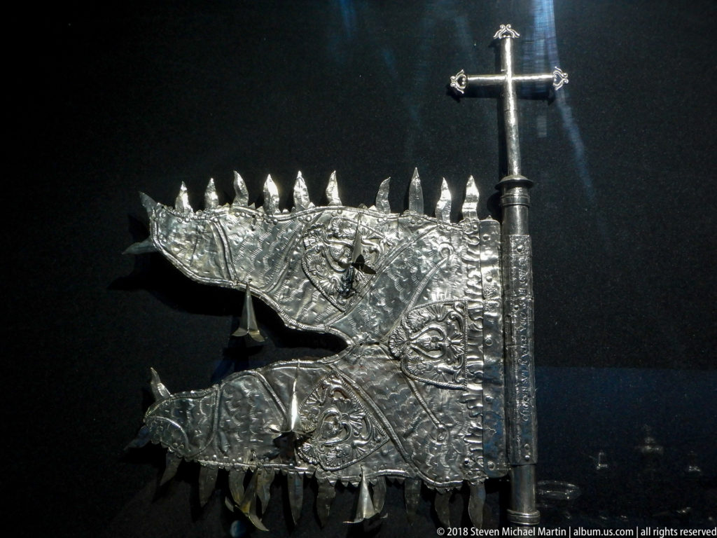 Silver-colored ceremonial standard with two embossed banner plates, pointed pendants, vertical staff, and cross finial.
