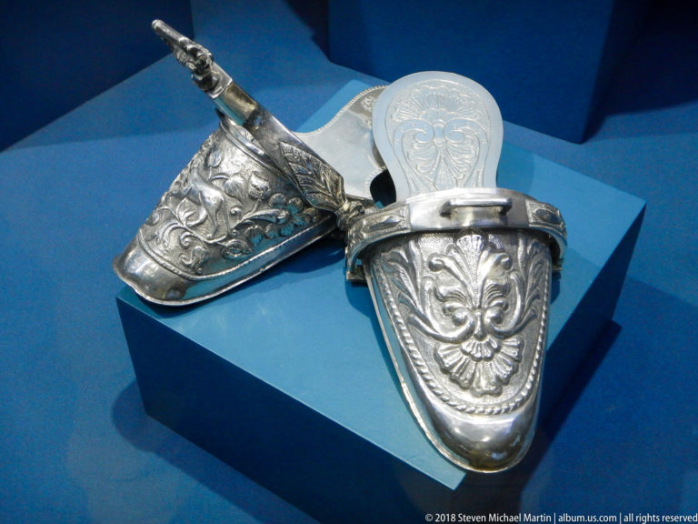 Pair of rigid silver-colored shoes covered with raised floral and scroll decoration on a blue museum plinth.