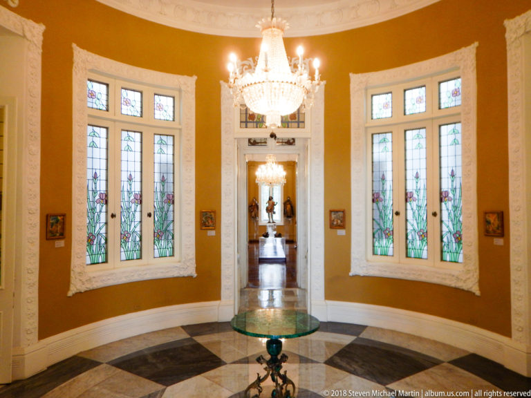 Curved ochre gallery with floral stained-glass windows, crystal chandelier, patterned floor, and a round display table.