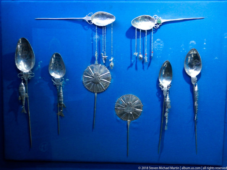 Silver spoons, ladles with hanging chains, and disc-headed pin-like ornaments arranged against a blue display background.