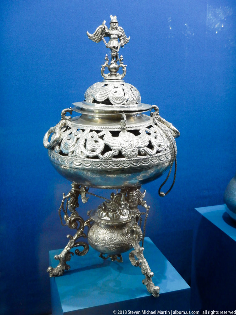 Ornate silver covered vessel with pierced openwork, sculptural finial, and scrolling three-legged base in a blue case.