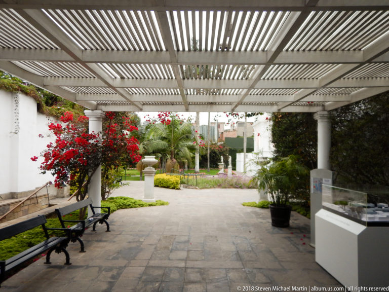 Slatted pergola over a paved museum garden walkway with benches, red bougainvillea, palms, lawn, and a glass display case.