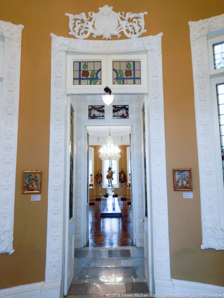 Aligned museum galleries seen through ornate white doorways, with chandeliers, stained-glass panels, paintings, and polished floors.