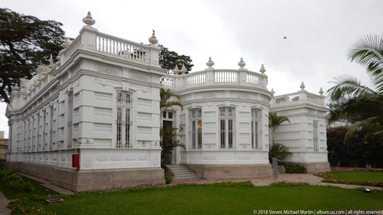 White Pedro de Osma Museum mansion seen from the garden, with rounded bay, tall windows, roof balustrades, and palms.