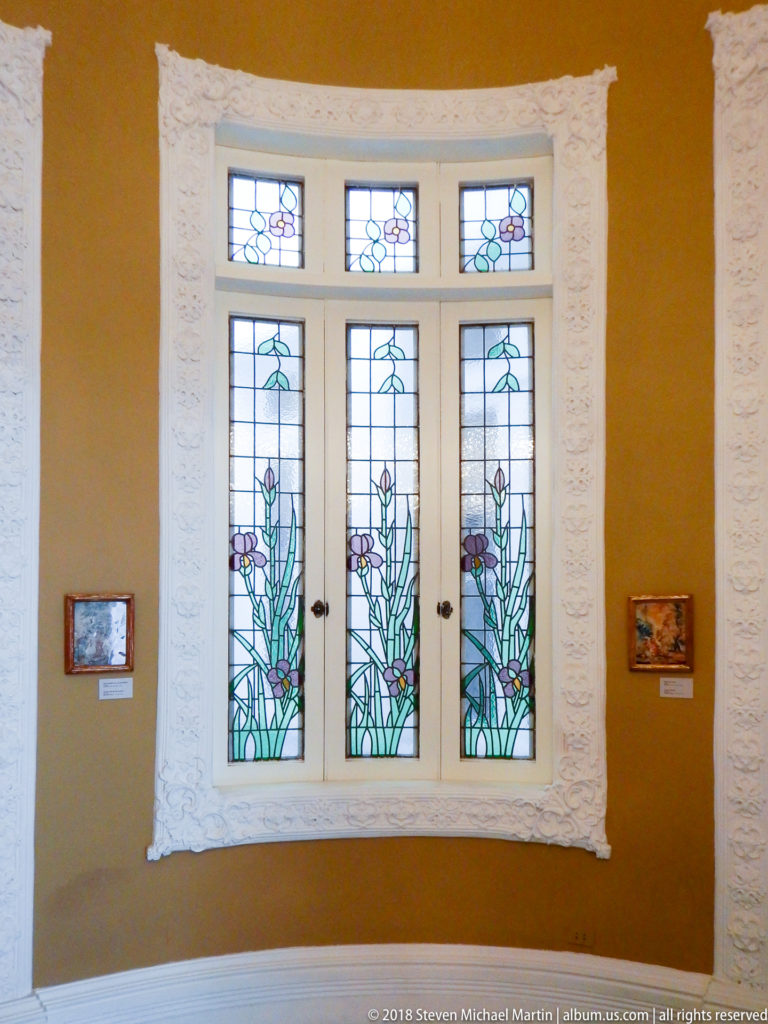 Three-panel stained-glass bay window with tall green floral motifs and purple accents, surrounded by ornate white plasterwork.