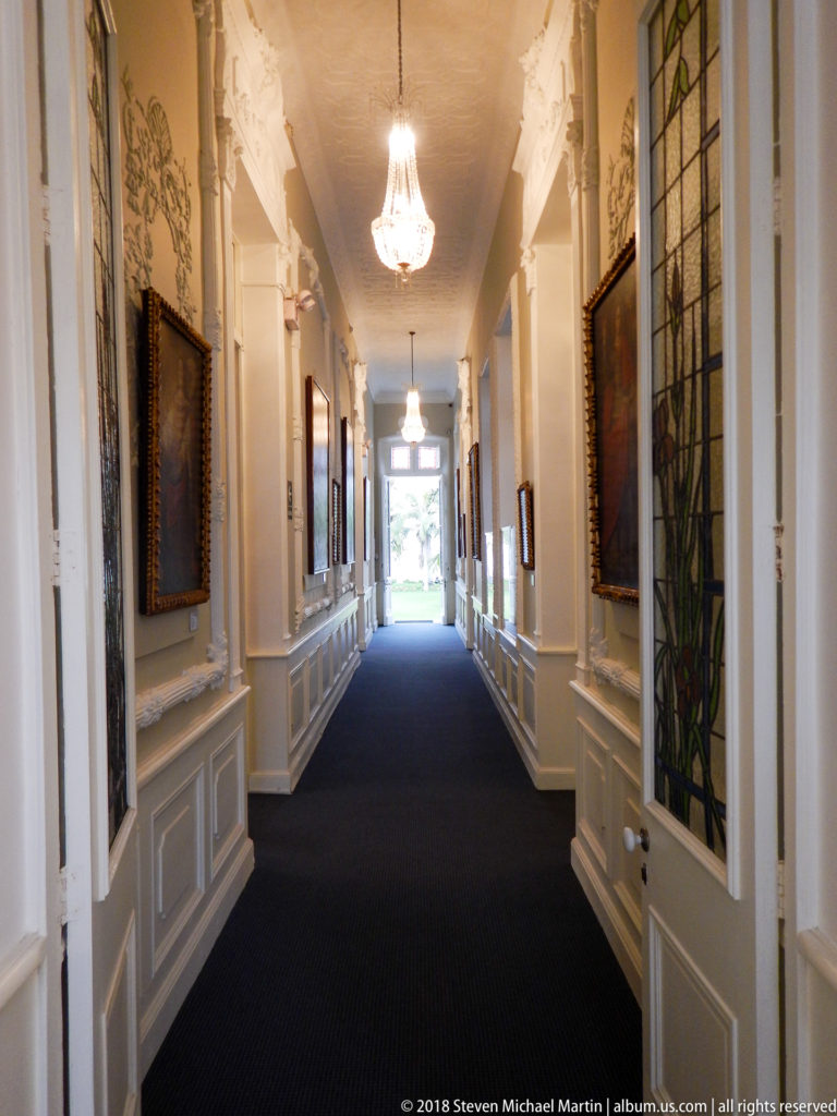 Long white-paneled museum corridor with dark runner, framed paintings, stained-glass panel, and hanging chandeliers.