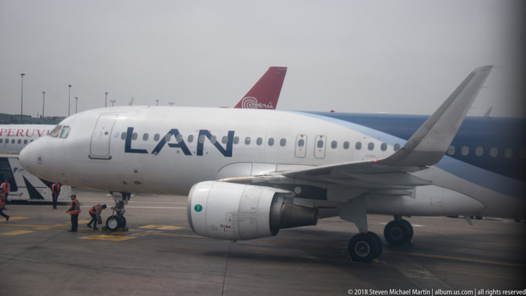 LAN passenger jet parked on an airport apron with ground crew and another aircraft behind.