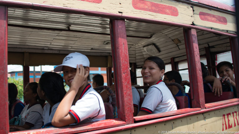 Students looking and smiling through the open windows of a worn red-and-cream bus.