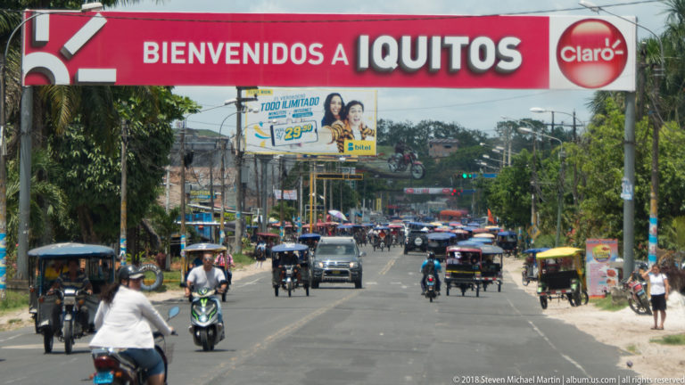 Red “Bienvenidos a Iquitos” banner spanning a busy road filled with mototaxis, motorcycles, cars, and pedestrians.