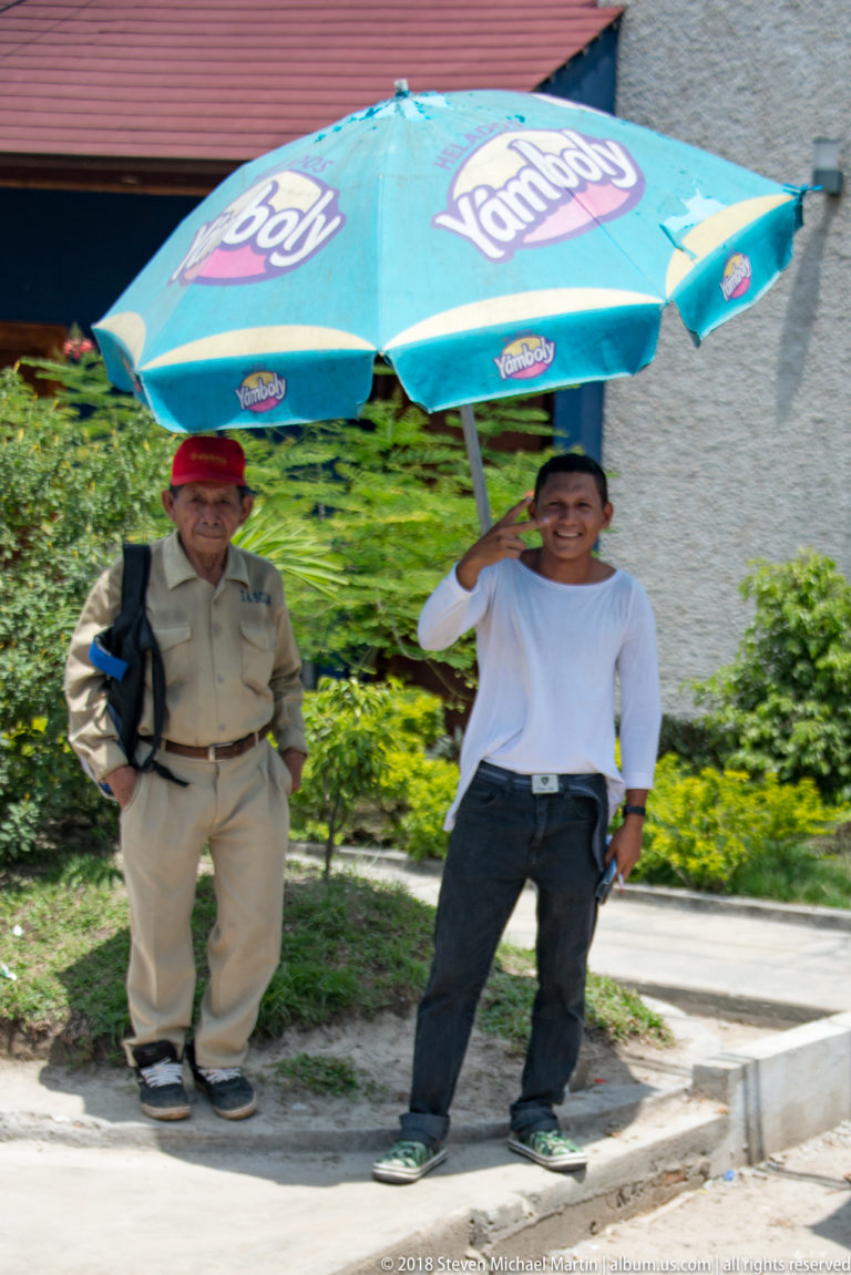 Two men standing beneath a turquoise advertising umbrella beside a curb, one in tan and one in white.